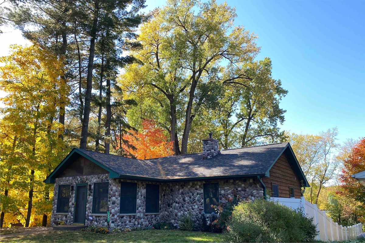The exterior of the home features a combination of stone and wood elements, set against a backdrop of vibrant autumn foliage. A chimney is visible on the roof. The surrounding trees contribute to a serene atmosphere, while a fence delineates the property.