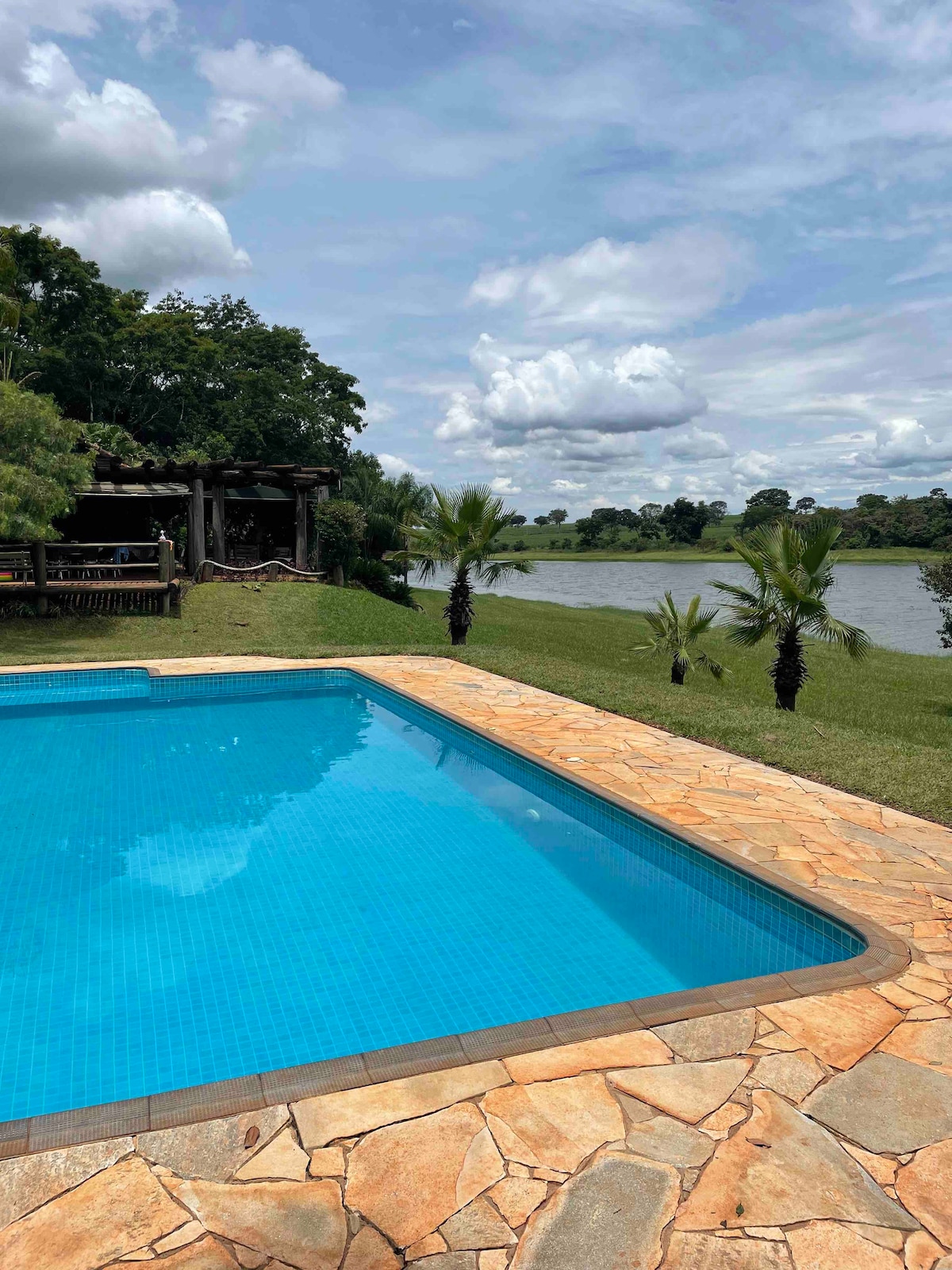 A clear swimming pool reflects the sky's blue hues, set within a stone patio. Lush green grass and small palm trees extend toward a serene river in the distance, providing a tranquil view of the natural surroundings.