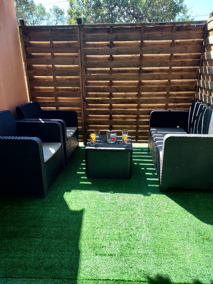 A comfortable outdoor seating area is presented, featuring four dark wicker chairs arranged around a small table. The space is enclosed by a wooden privacy wall, with artificial grass covering the ground, creating a relaxing environment for guests.
