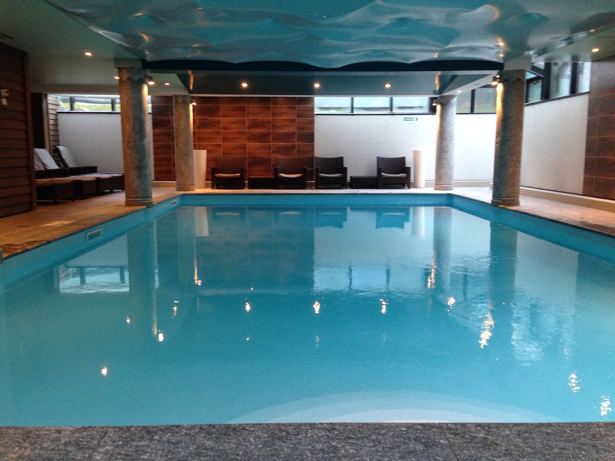 An indoor swimming pool is surrounded by sleek stone columns. The water's surface glistens in bright turquoise, reflecting the soft lighting above. Comfortable lounge chairs are positioned along one side of the pool, creating a relaxing atmosphere for guests.