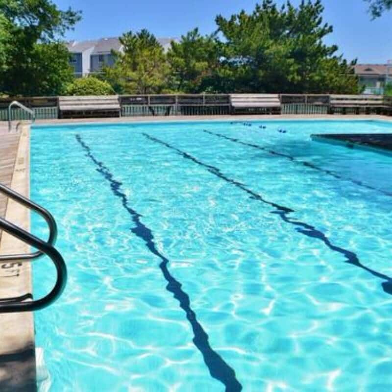 A clear blue community pool is visible, reflecting sunlight on its surface. Surrounding the pool are wooden lounge chairs arranged along the deck. Trees and greenery provide a natural backdrop, contributing to an inviting recreational area.