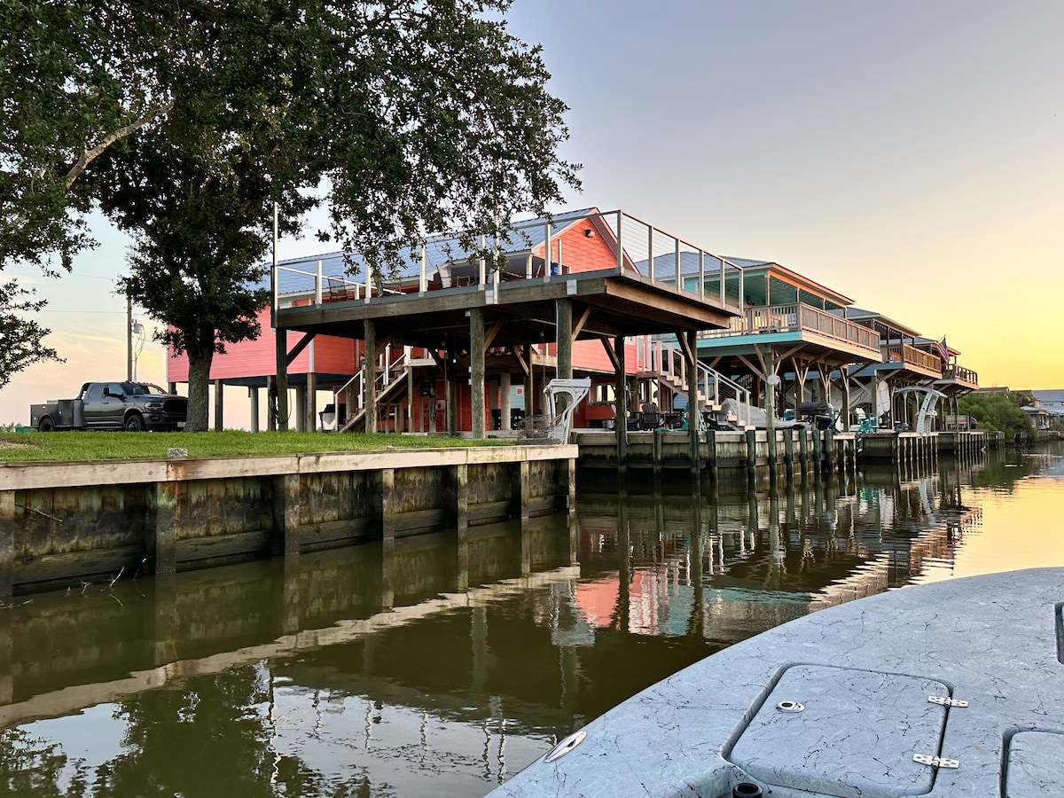 A view of a multi-level camp house along a private canal is depicted. The structure features a spacious deck over a boat slip, with vibrant colours on the exterior. Calm waters reflect the surroundings, while boats are moored beneath the wooden framework.