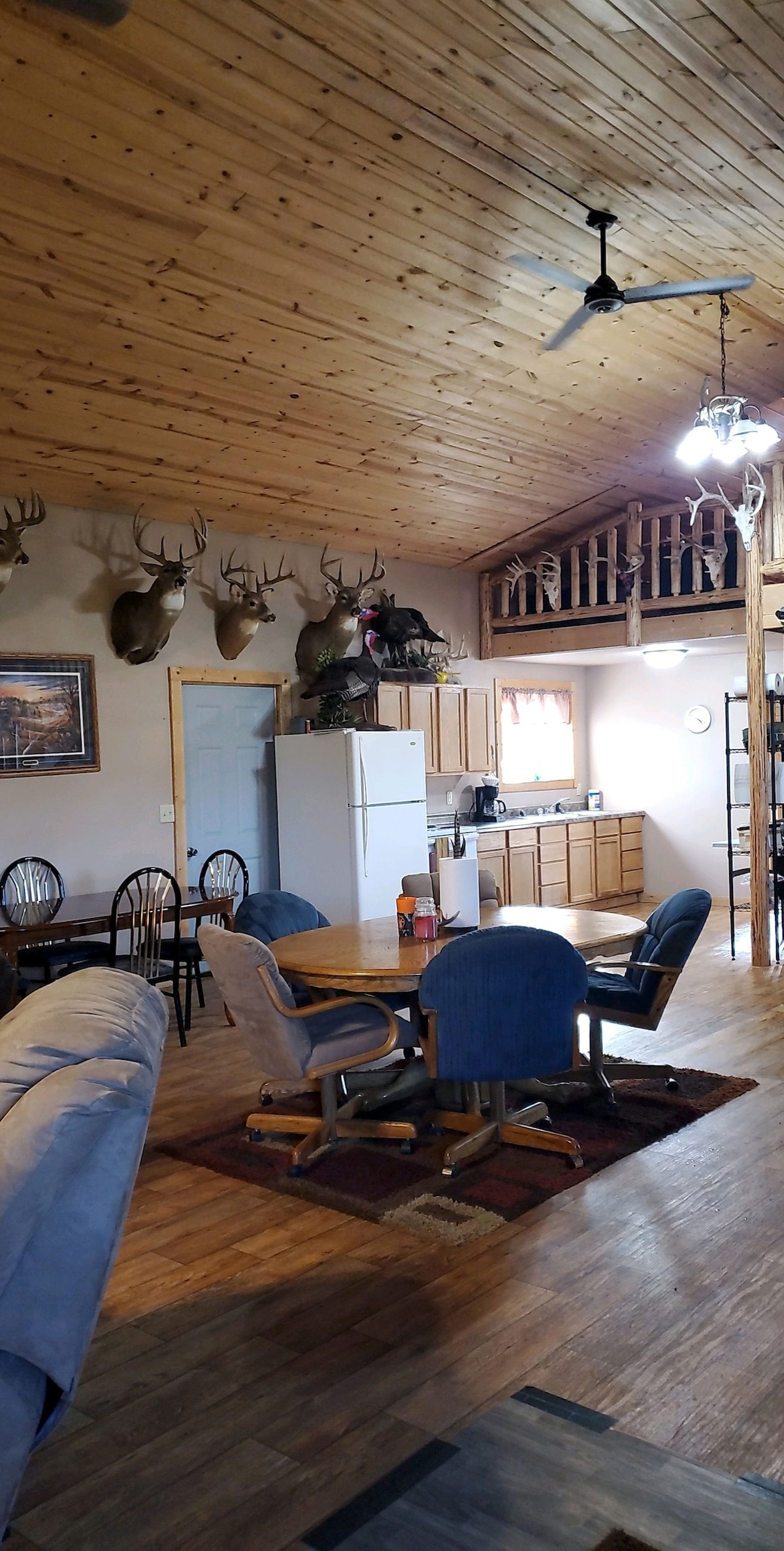 A spacious dining area features a wooden table surrounded by six chairs. A refrigerator is visible against the wall, with a fully equipped kitchen seen in the background. Mounted trophies adorn the walls, and natural wood paneling enhances the rustic charm of the lodge.