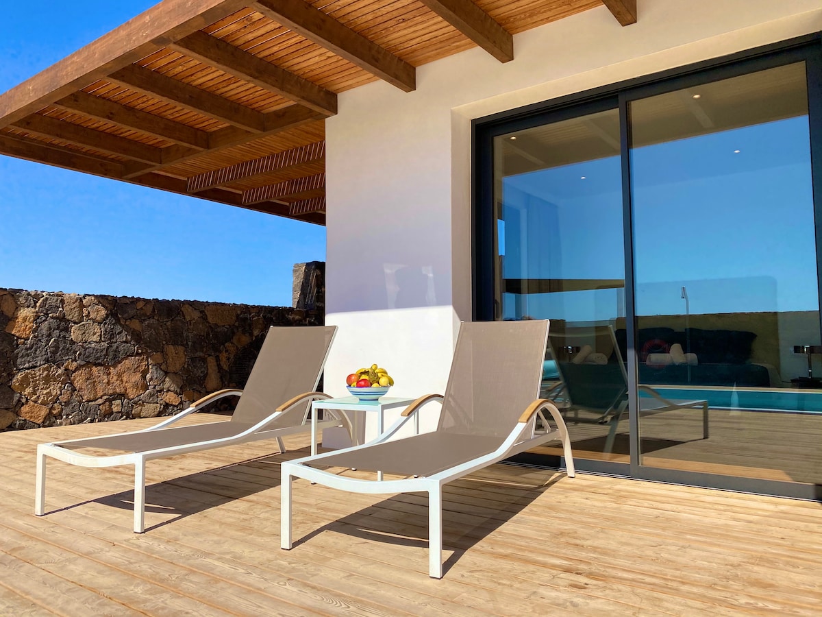 Two lounge chairs are positioned on a wooden terrace, while a small table holds a bowl of fresh fruits. Large glass doors provide a view of the interior, with natural light flooding the space and the backdrop of mountains visible beyond the patio.