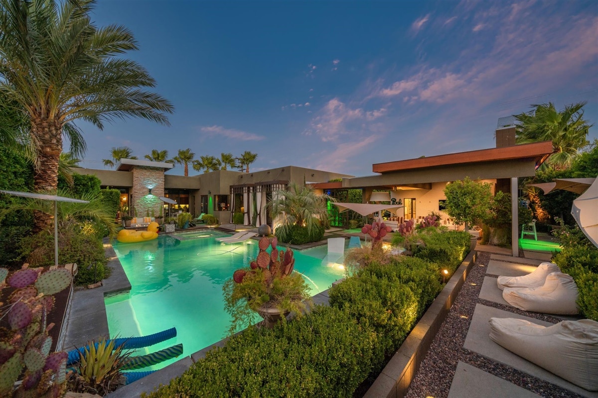 A serene outdoor space features a spacious salt-water pool surrounded by lush landscaping, including palm trees and colorful cacti. Comfortable loungers are arranged beside the pool, creating an inviting area for relaxation. The evening sky is painted with soft hues, enhancing the tranquil atmosphere.