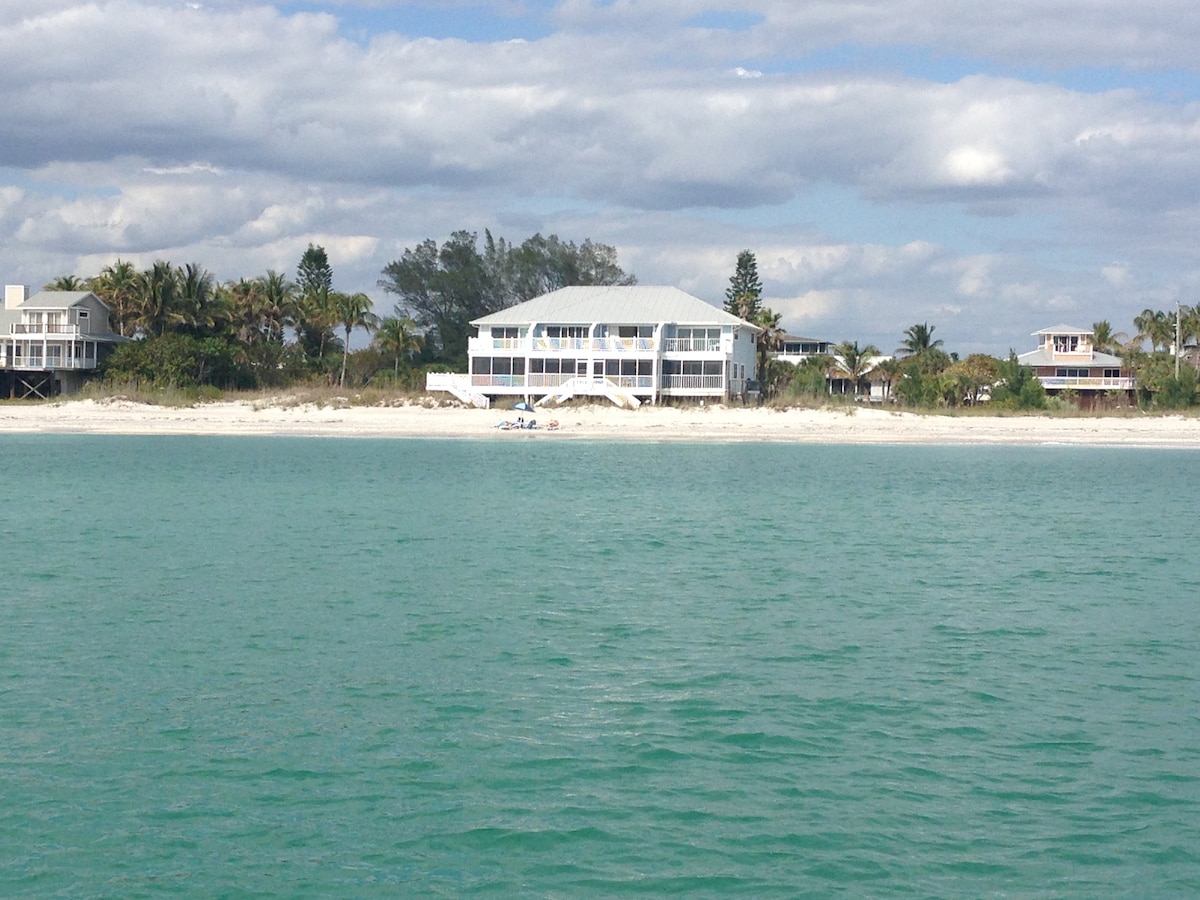 A two-story beach house is viewed from the water, with a white exterior and large balconies overlooking the shoreline. Lush palm trees surround the property, and soft white sand beaches are visible in the foreground, against a backdrop of gentle waves and a cloudy sky.