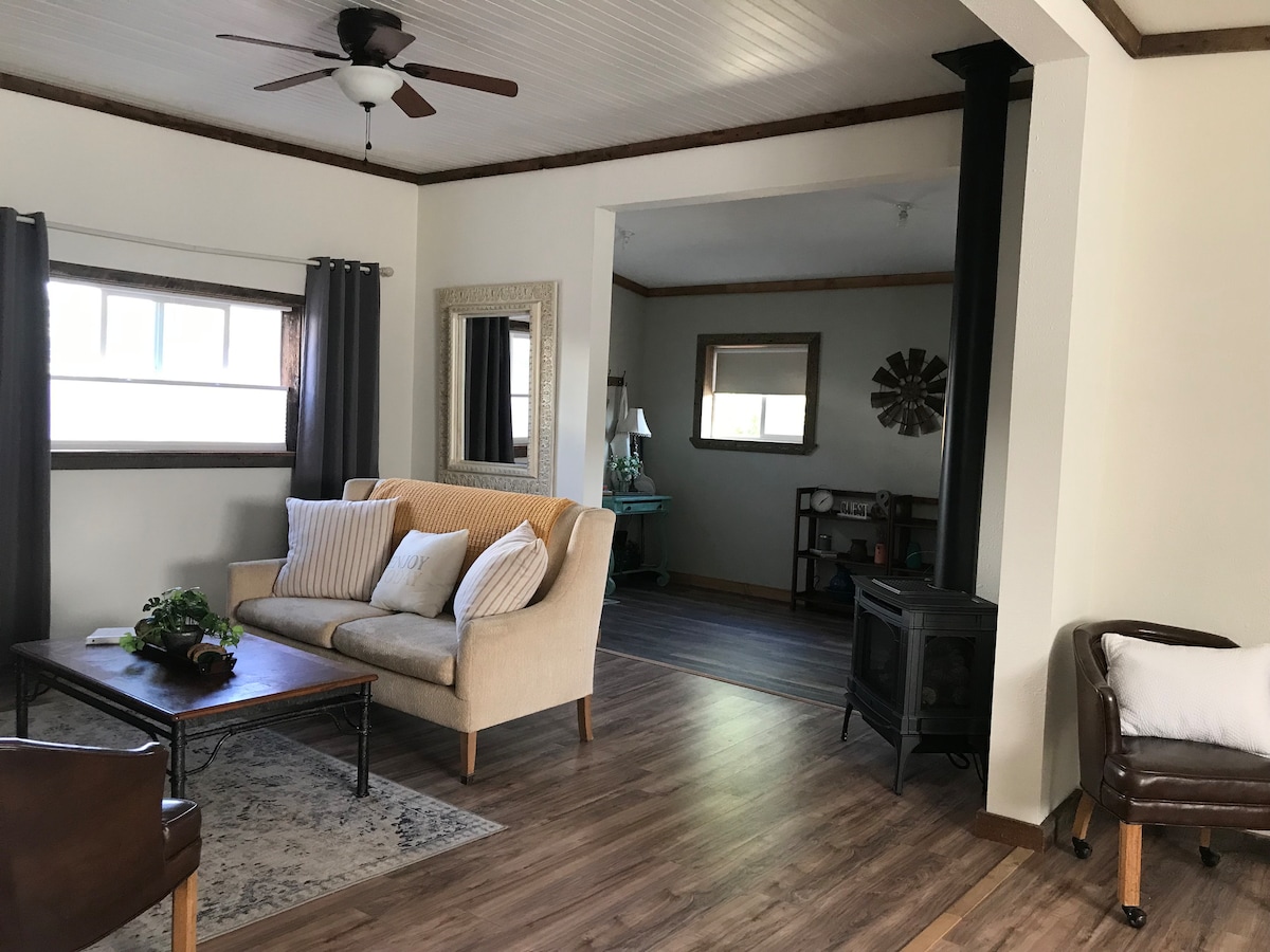 A spacious living area is illuminated by natural light from two windows. A comfortable sofa with decorative pillows faces a wooden coffee table. Nearby, a small side table and a wall-mounted mirror reflect the room's decor. A metal fireplace adds warmth to the space.