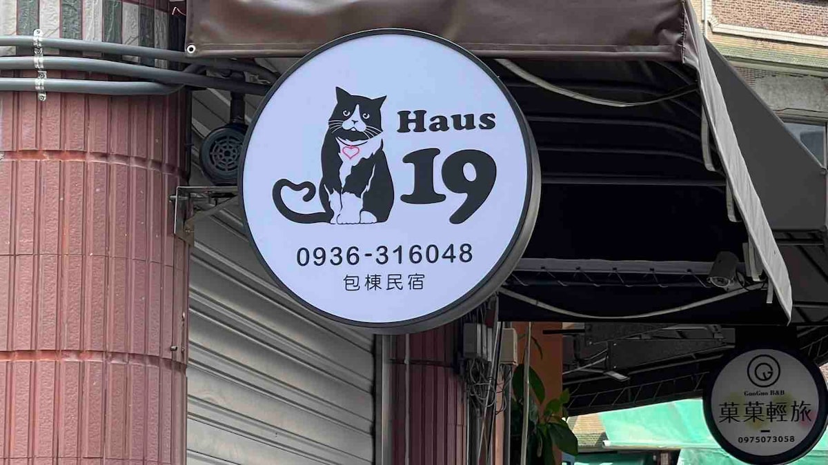 A round sign displaying the name 'Haus 19' is featured, accompanied by an illustration of a cat. The sign includes a phone number for contact and is mounted above a storefront, partially shaded by an awning.
