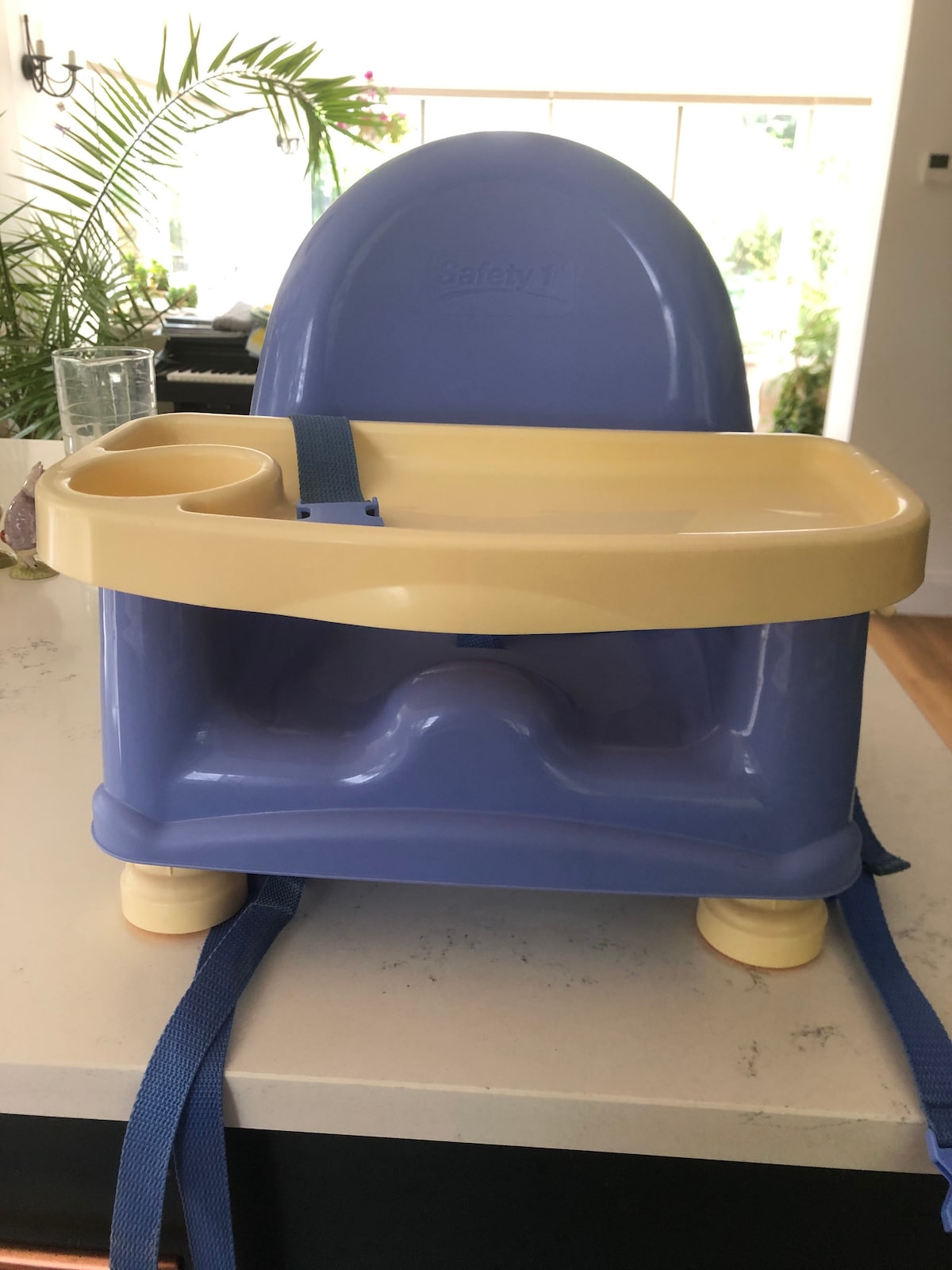 A child-sized high chair, featuring a light blue and yellow design. The chair includes a food tray with a cup holder and safety straps for secure seating. The sturdy base is composed of four legs with yellow feet, resting on a countertop in a bright and airy environment.