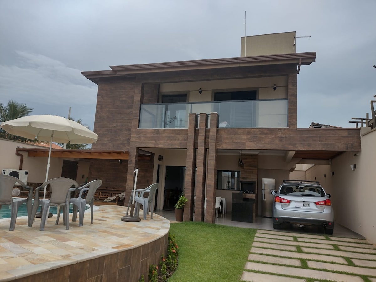 The exterior of the property features a two-story structure with a contemporary design, complemented by large windows and a spacious balcony. A car is parked in the driveway next to well-maintained grass and a stone pathway. Poolside seating is visible under an umbrella.