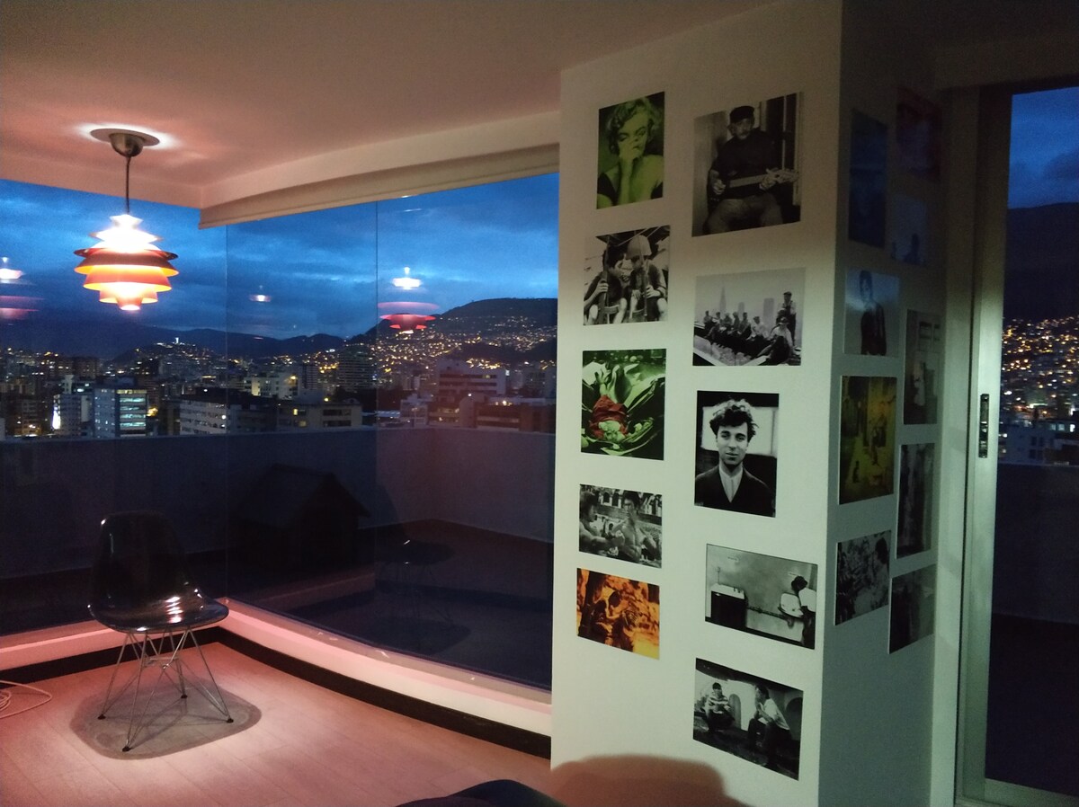 An artistic corner of the penthouse features a sleek black chair beside floor-to-ceiling glass windows, offering views of the cityscape at dusk. A wall adorned with various photographs adds character, while warm pendant lighting creates a relaxed ambiance.