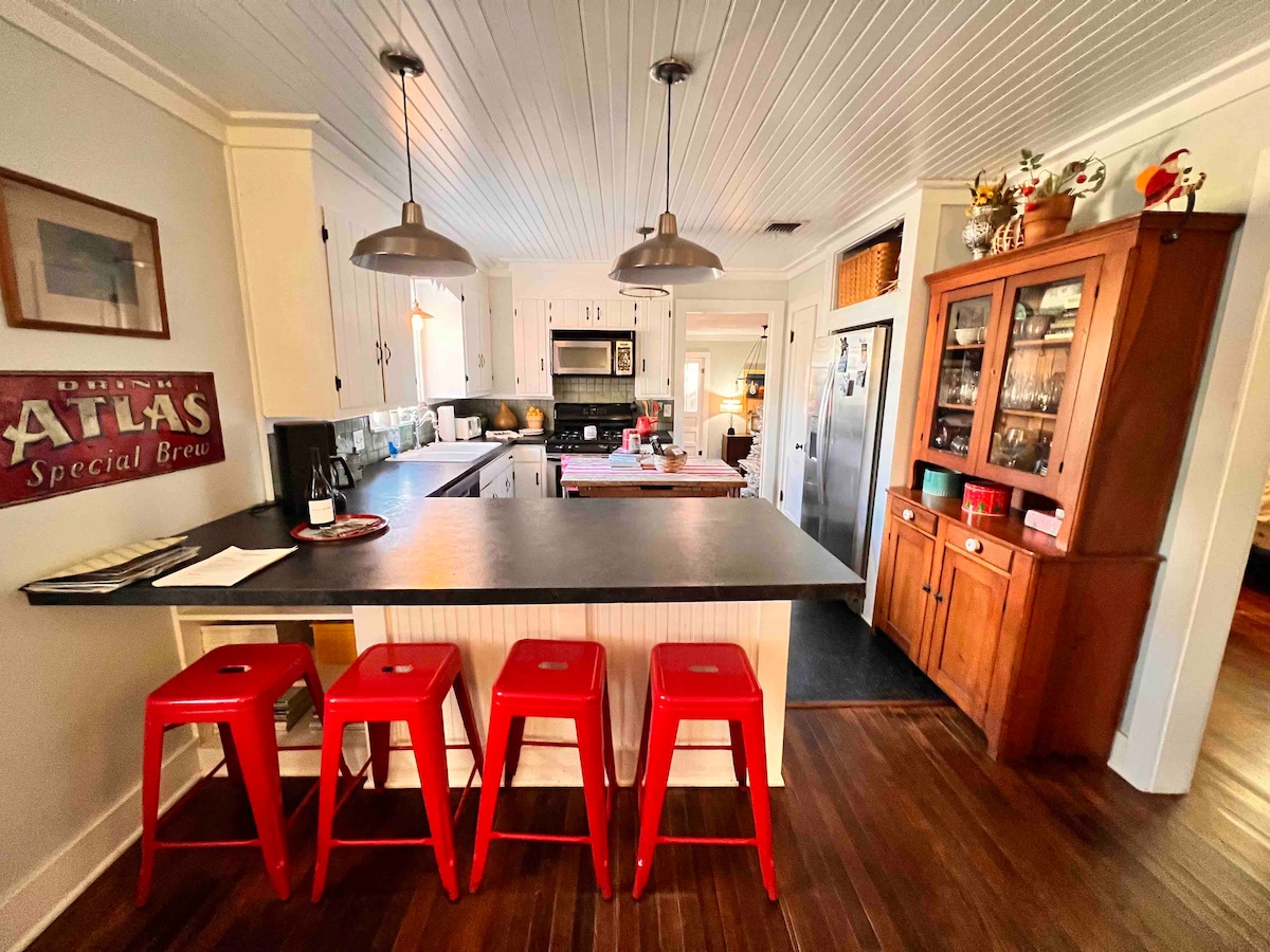 A kitchen space features a central island with a dark countertop and red stools arranged along one side. Stainless steel appliances are present, along with wooden cabinetry. Natural light fills the area, enhancing the warm tones of the wood flooring.
