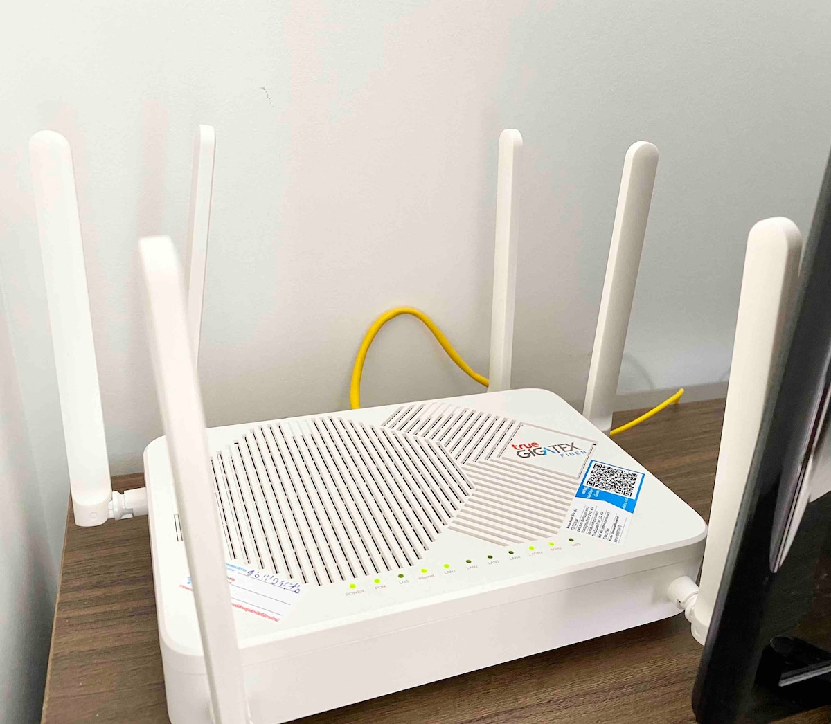 A modern Wi-Fi router is placed on a wooden surface, featuring multiple antennas for optimal connectivity. The router's lights indicate its operational status, while a yellow Ethernet cable connects it to the wall, ensuring reliable internet access for guests.