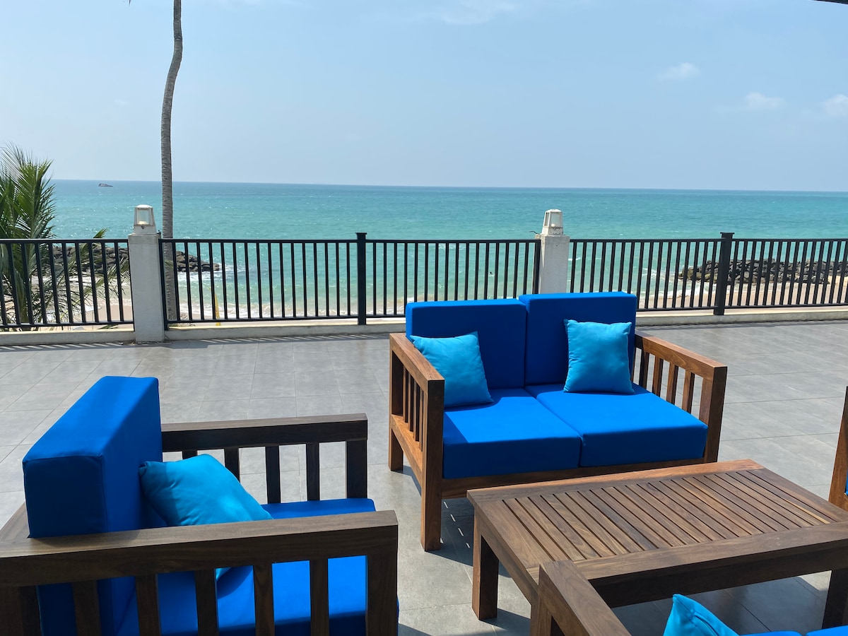 An outdoor seating area features modern blue sofas with matching cushions, arranged around a wooden coffee table. The space offers a clear view of the calm turquoise ocean, with a railing bordering the terrace, enhancing the coastal ambiance.