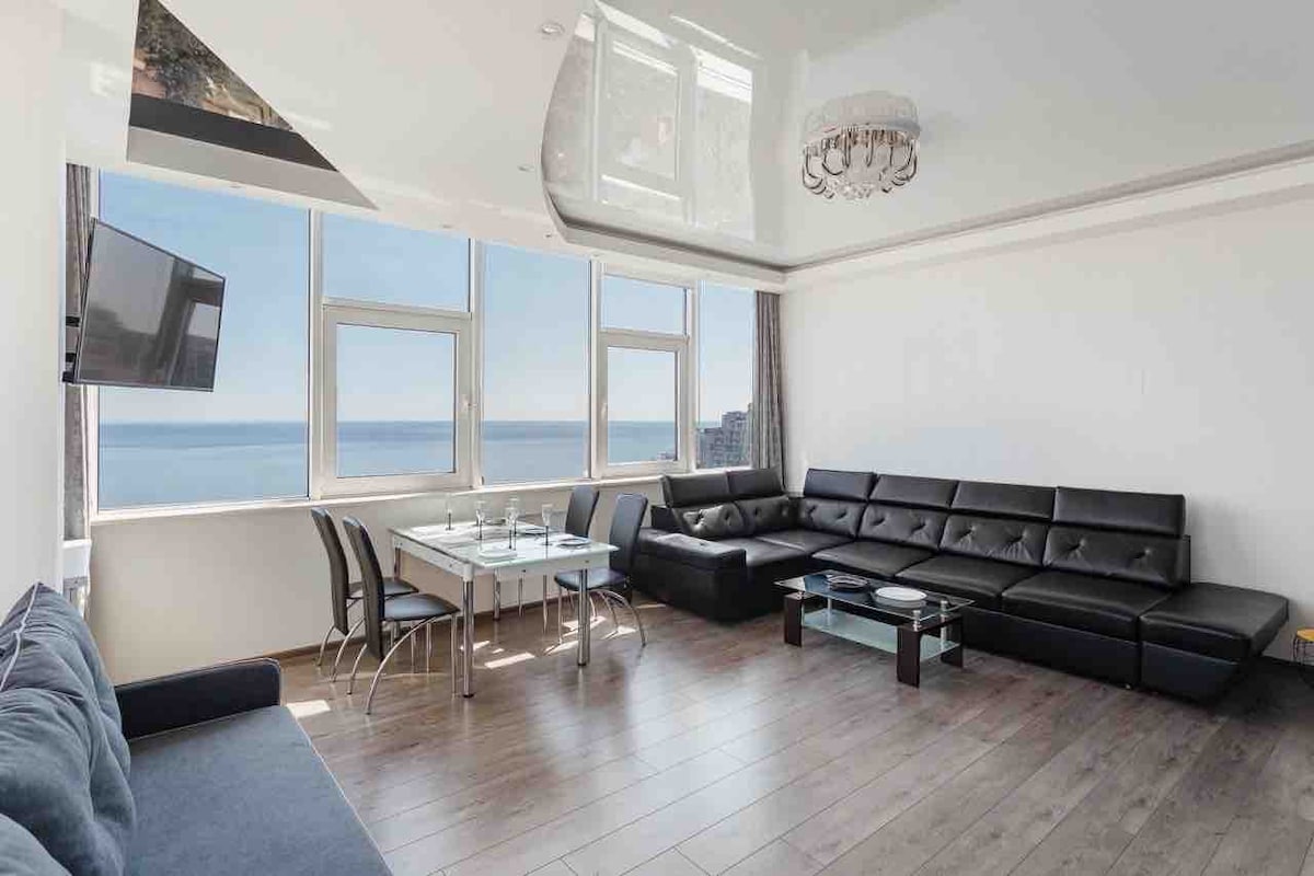 A modern living area features large windows offering a view of the sea. A sleek black sectional sofa and a glass coffee table provide comfortable seating. A dining table with metallic chairs complements the space, while a flat-screen TV is mounted on the wall.
