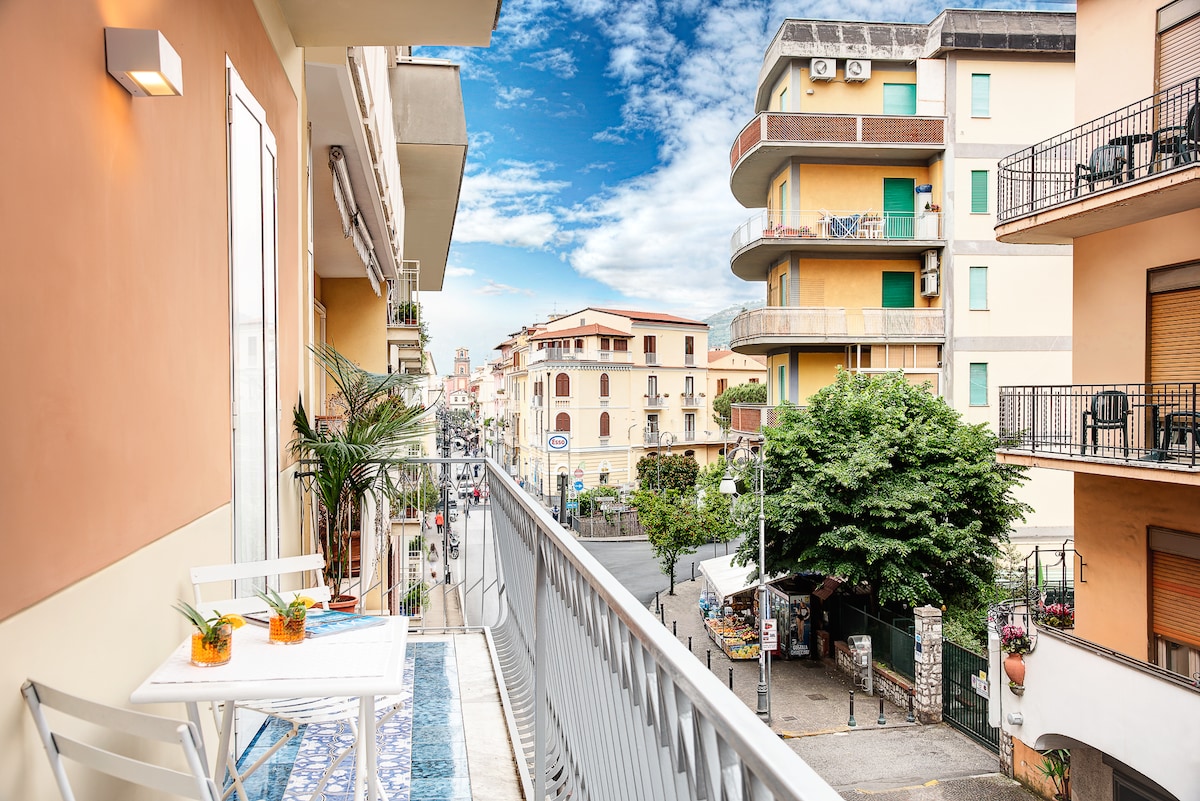 A wraparound terrace is visible, featuring a small outdoor table with seating for two. The view includes the vibrant street below adorned with trees, buildings, and various shops. Clear skies with scattered clouds create a bright atmosphere, highlighting the lively surroundings.