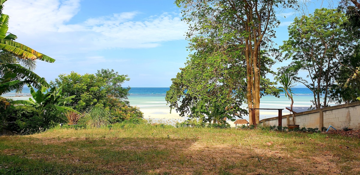 A lush, green beachfront yard is visible, featuring a clear view of the calm sea and a sandy shore. Various trees frame the scene, offering natural shade. The sky showcases a blend of soft clouds and sunlight, adding to the peaceful coastal ambiance.