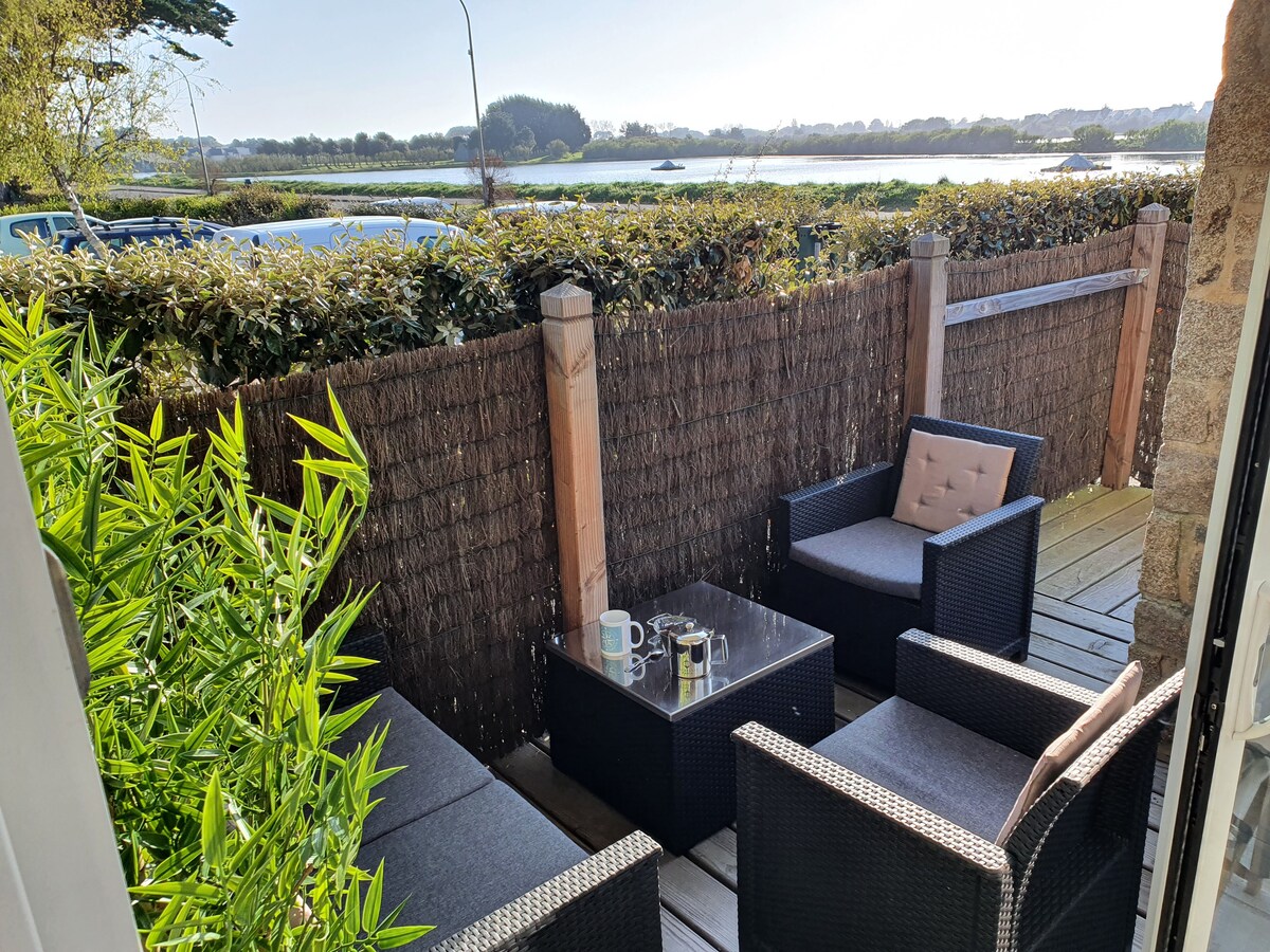 An outdoor terrace features a cozy sitting area with two armchairs and a small coffee table. Lush green plants surround the space, providing privacy. A scenic view of a river and distant landscape is visible in the background, enhancing the tranquil atmosphere.