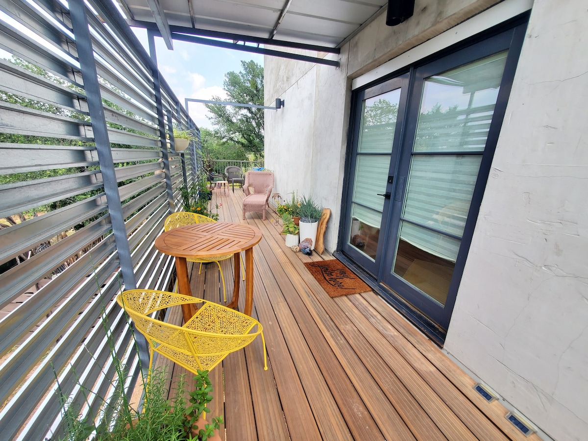 A spacious upper deck is presented with wooden flooring and metal railing, featuring a round wooden table accompanied by two vibrant yellow chairs. Potted plants add greenery to the space, while a cozy seating area is visible, inviting relaxation amidst natural surroundings.