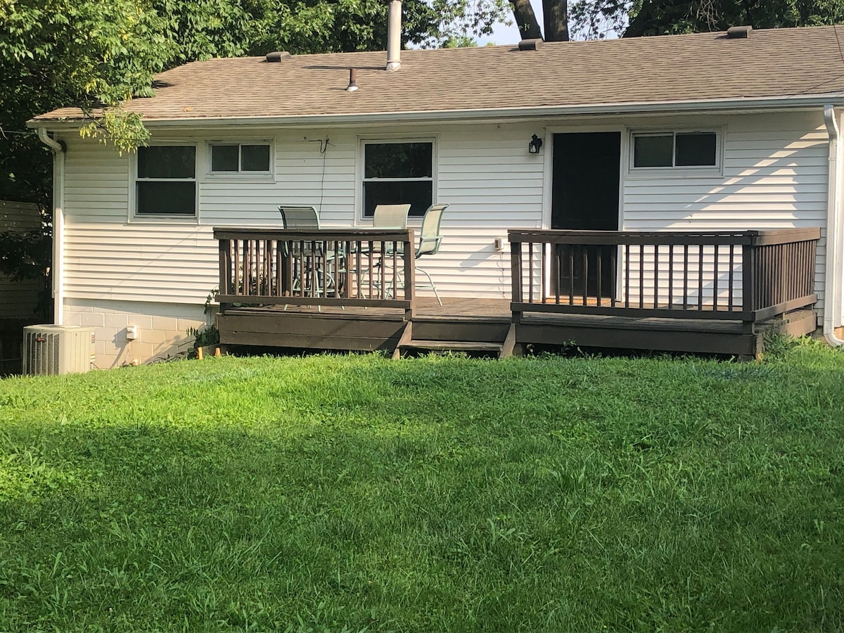 A single-story home is set against a backdrop of green grass. A wooden deck extends from the house, featuring a small table and four chairs. Windows are placed symmetrically on either side of the entrance door, providing natural light.