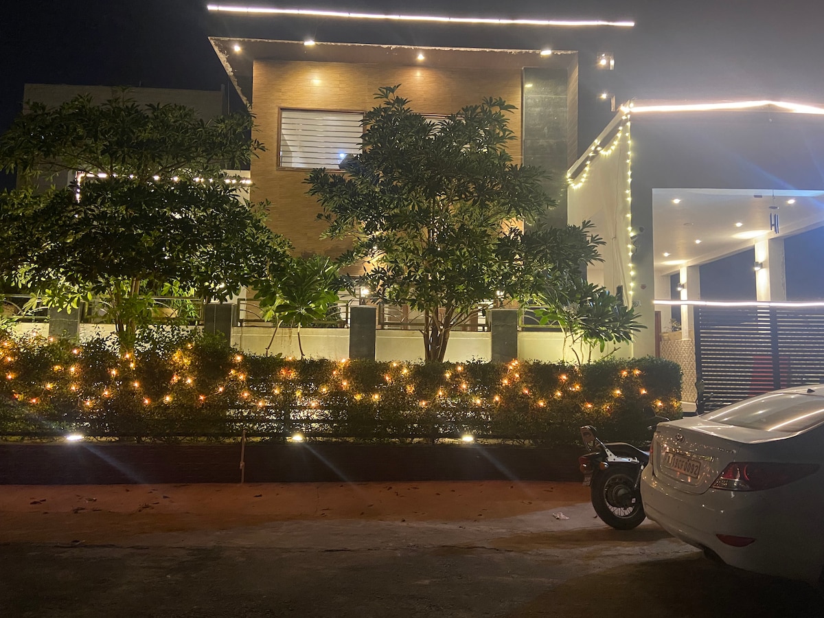 The villa is illuminated at night, showcasing modern architecture with wooden accents and lush greenery surrounding the entrance. Soft lights are placed along the garden and pathway, enhancing the welcoming ambiance. A parked car is visible on the driveway near the property.