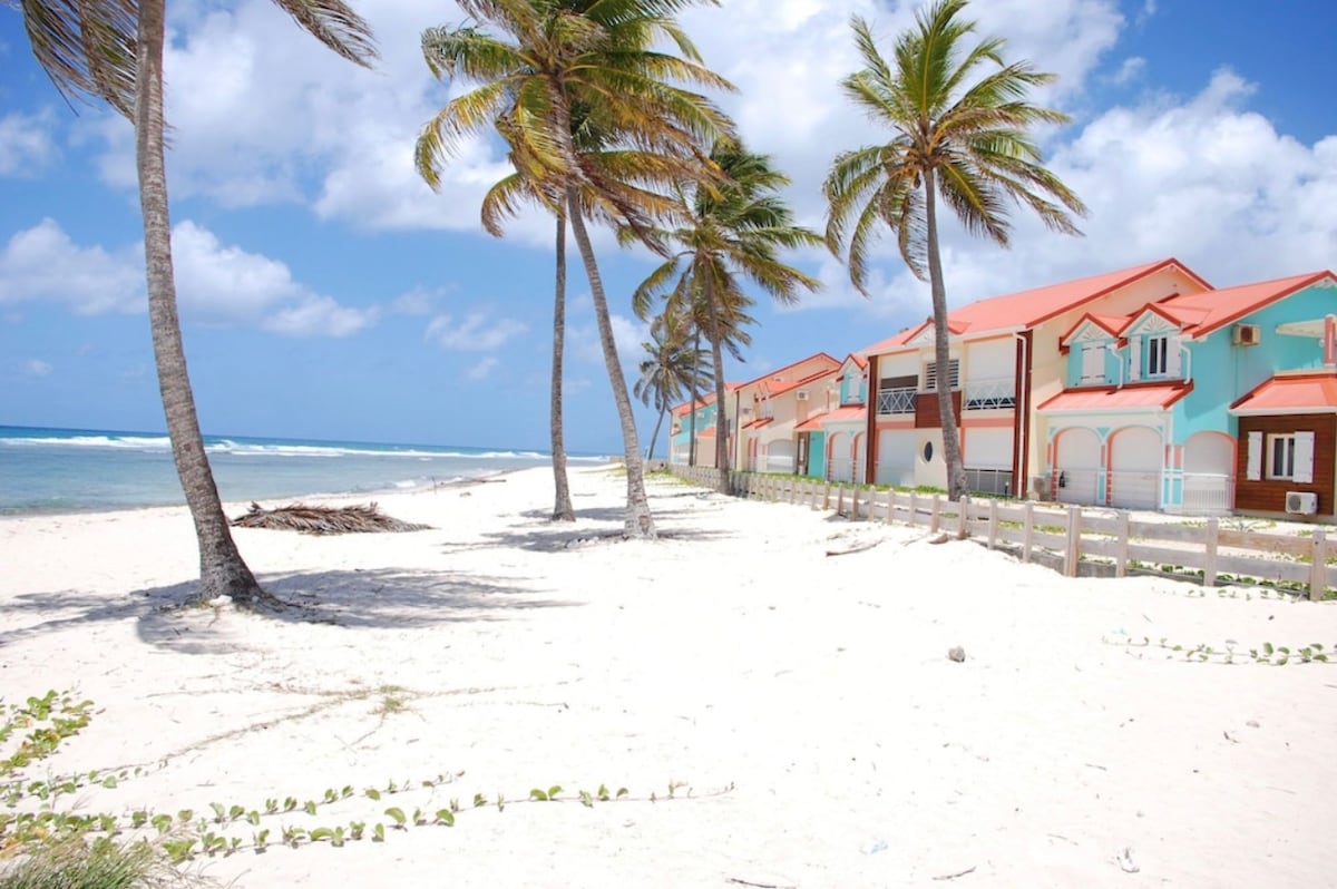 A beachfront view showcases a series of modern duplex villas with vibrant colors and red roofs. Tall palm trees line the sandy shore, with gentle waves visible in the distance. The serene atmosphere is complemented by a clear blue sky with scattered clouds.