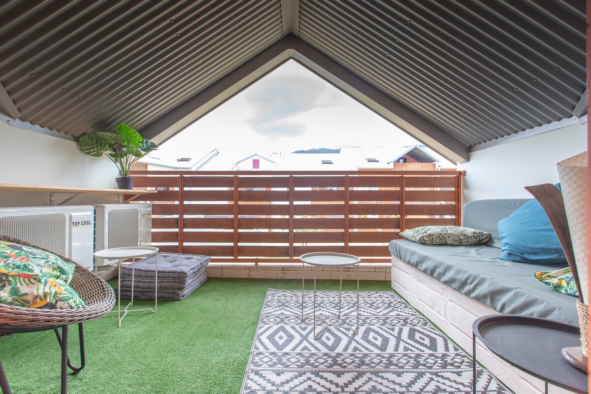 A covered terrace is presented with a cozy seating arrangement, featuring a spacious couch, decorative cushions, and small tables. A patterned rug adds texture to the green flooring. Natural light filters through the angled roof, enhancing the inviting atmosphere.