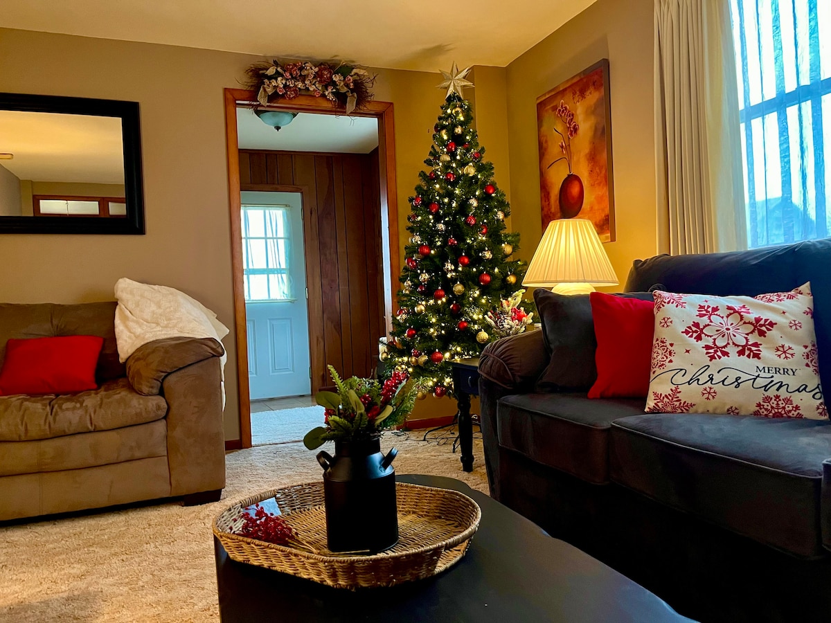 A cozy living room is presented, featuring a decorated Christmas tree with twinkling lights and colorful ornaments. A brown couch and a dark sofa are arranged around a coffee table adorned with a decorative tray. Seasonal decor is visible, contributing to a festive atmosphere.