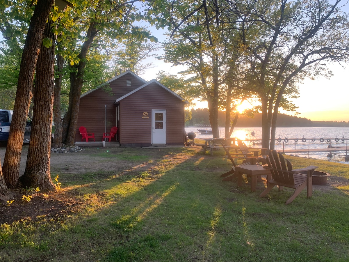 A rustic cabin is nestled among trees, with a grassy area in front featuring wooden seating. The sun sets beyond the nearby lake, casting warm light on the cabin's exterior, while a small beach area and dock are visible in the background.