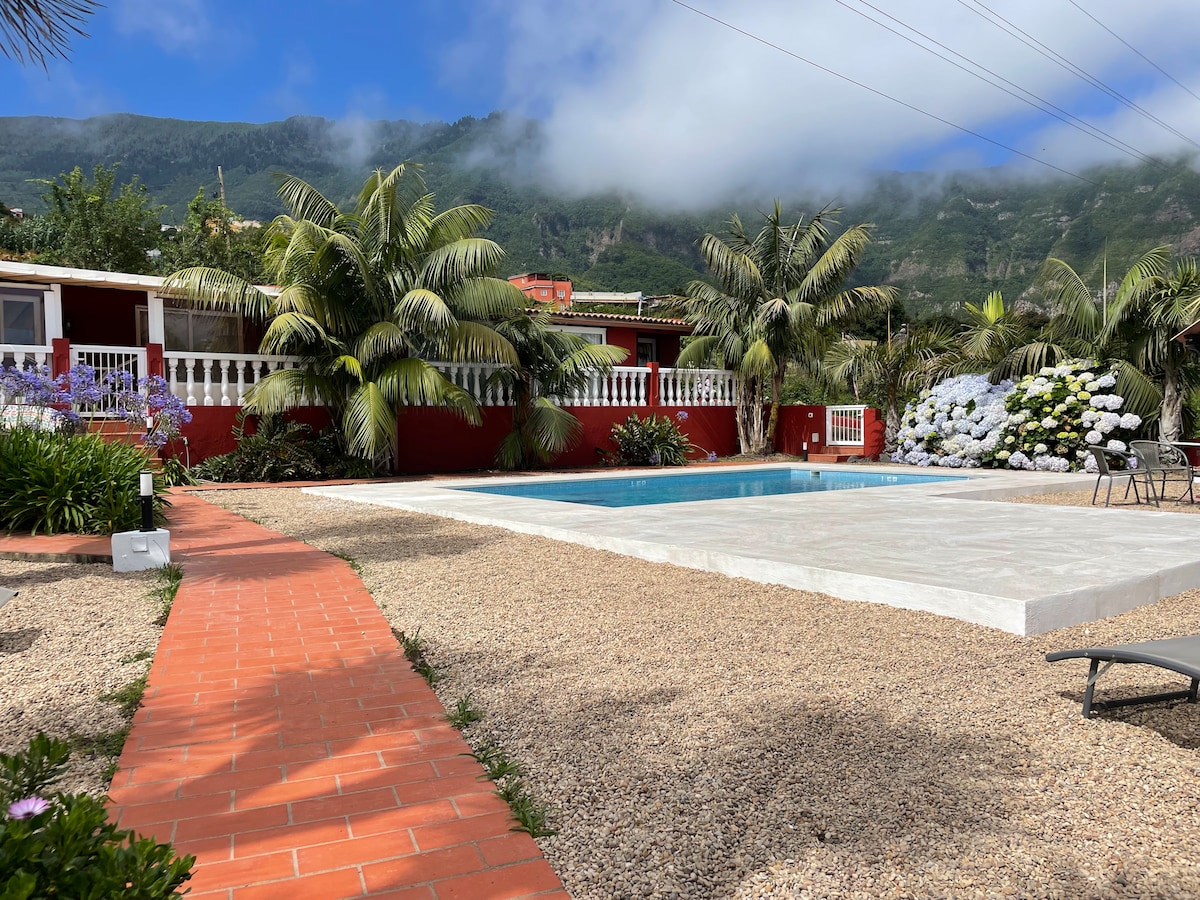 A well-maintained garden area features a large swimming pool surrounded by palm trees and blooming flowers. A brick pathway leads through the gravel landscape, enhancing the serene rural setting with mountains in the background and walls painted in a warm red hue.