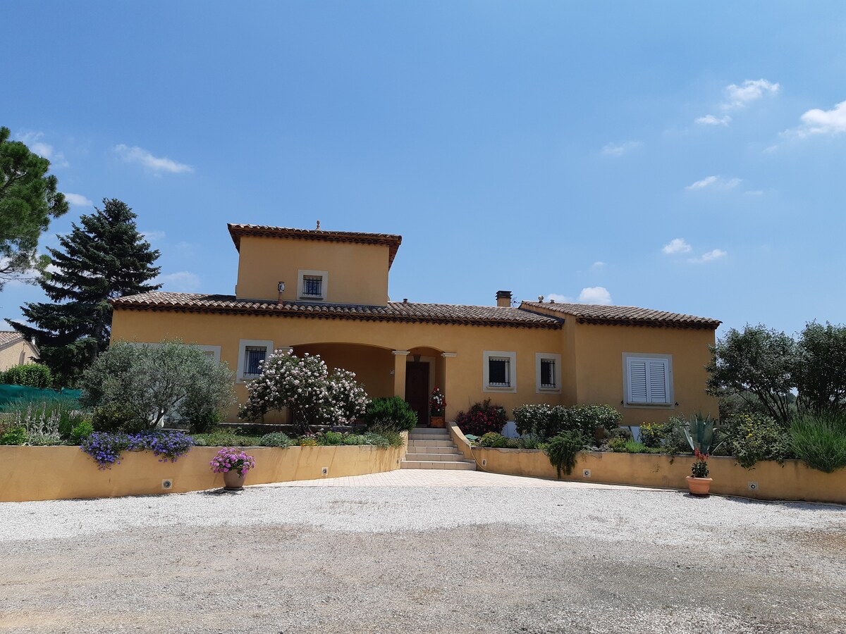 A spacious villa is presented, showcasing a warm yellow facade and a tiled roof. Surrounding greenery includes olive trees and flower beds, enhancing the welcoming entrance. A gravel driveway leads to the front steps, framed by pots of vibrant flowers.