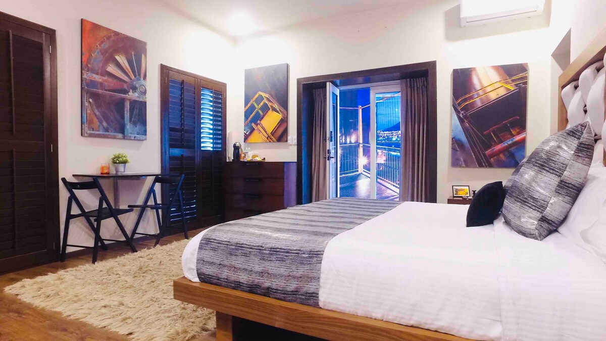 A spacious bedroom features a large bed with a neatly arranged coverlet. Two artworks adorn the walls, and a small table is positioned in the corner. Light streams in from the sliding glass doors, which open to a view of the city skyline.