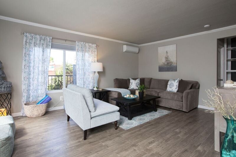 A living area features a comfortable gray sofa and an accent chair, arranged around a dark coffee table. Natural light enters through a window adorned with floral curtains. A neutral color palette is complemented by decorative accents, creating a welcoming space for relaxation.