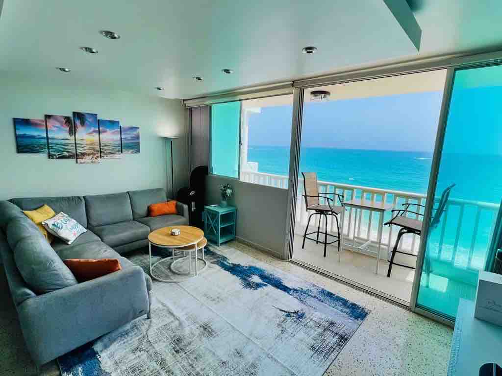 A modern living area features a large gray sofa with colorful pillows and a round coffee table. A vibrant rug adds texture, while a stunning ocean view is visible through expansive sliding glass doors leading to the balcony.