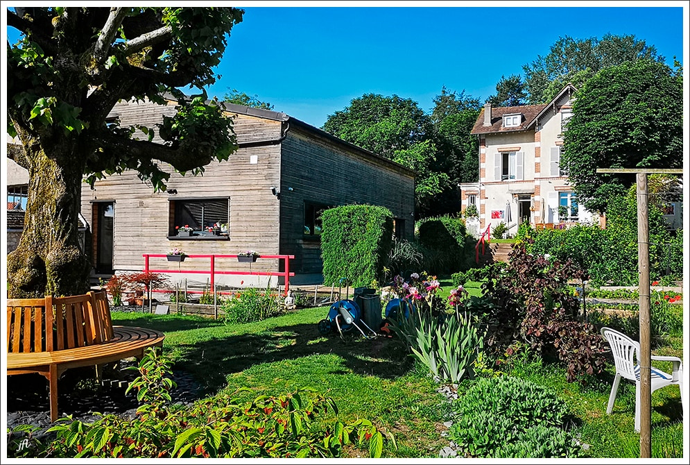 A wooden house with a sloped roof is surrounded by a lush garden, featuring colorful flowers and greenery. The scene includes a bench and seating area, providing a serene atmosphere. The property is adjacent to another traditional house, enhancing the peaceful setting.