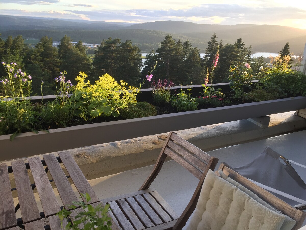 A balcony area is surrounded by lush planters filled with blooming flowers, offering a vibrant view of the distant hills and water. Wooden chairs and a table provide space for relaxation, with the scenic landscape creating a serene backdrop.