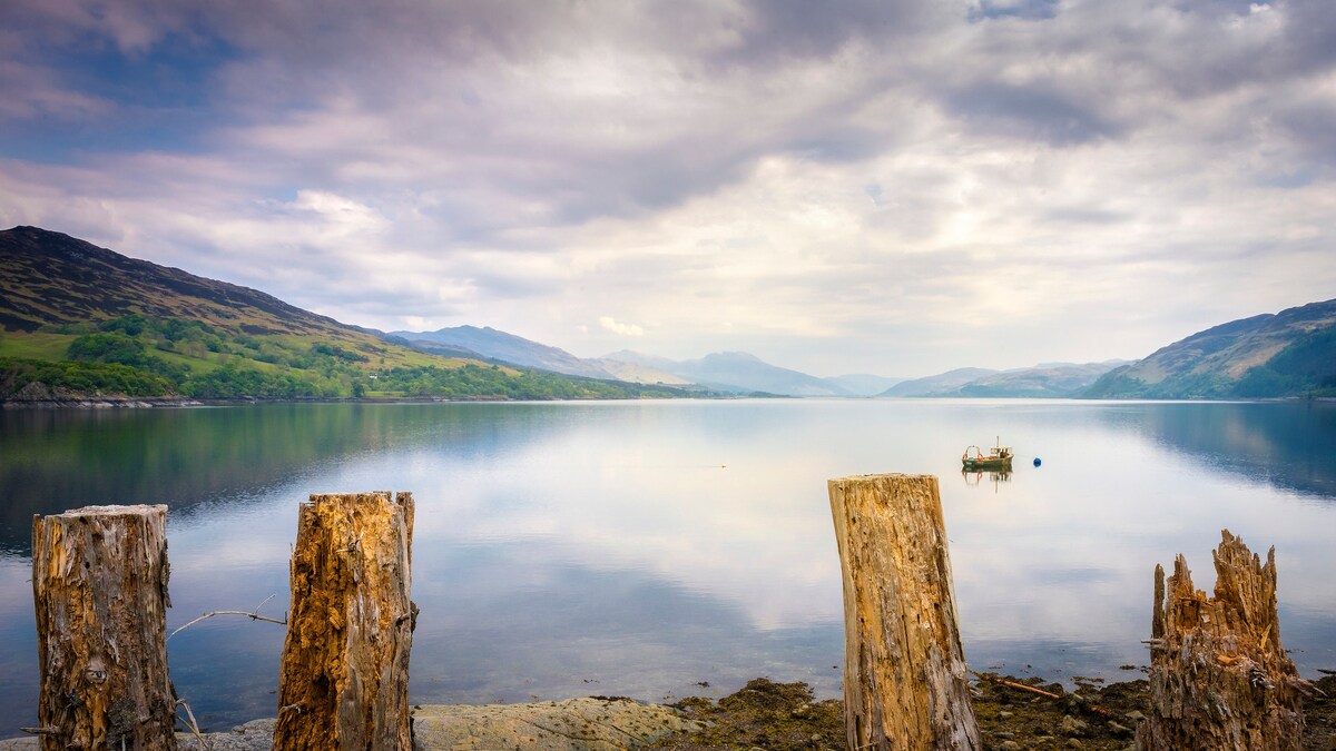A serene view of Loch Carron reflects the surrounding hills under a cloudy sky. Stumps of weathered logs stand in the foreground, leading the eye to a small boat anchored on the calm water, creating a sense of tranquility.