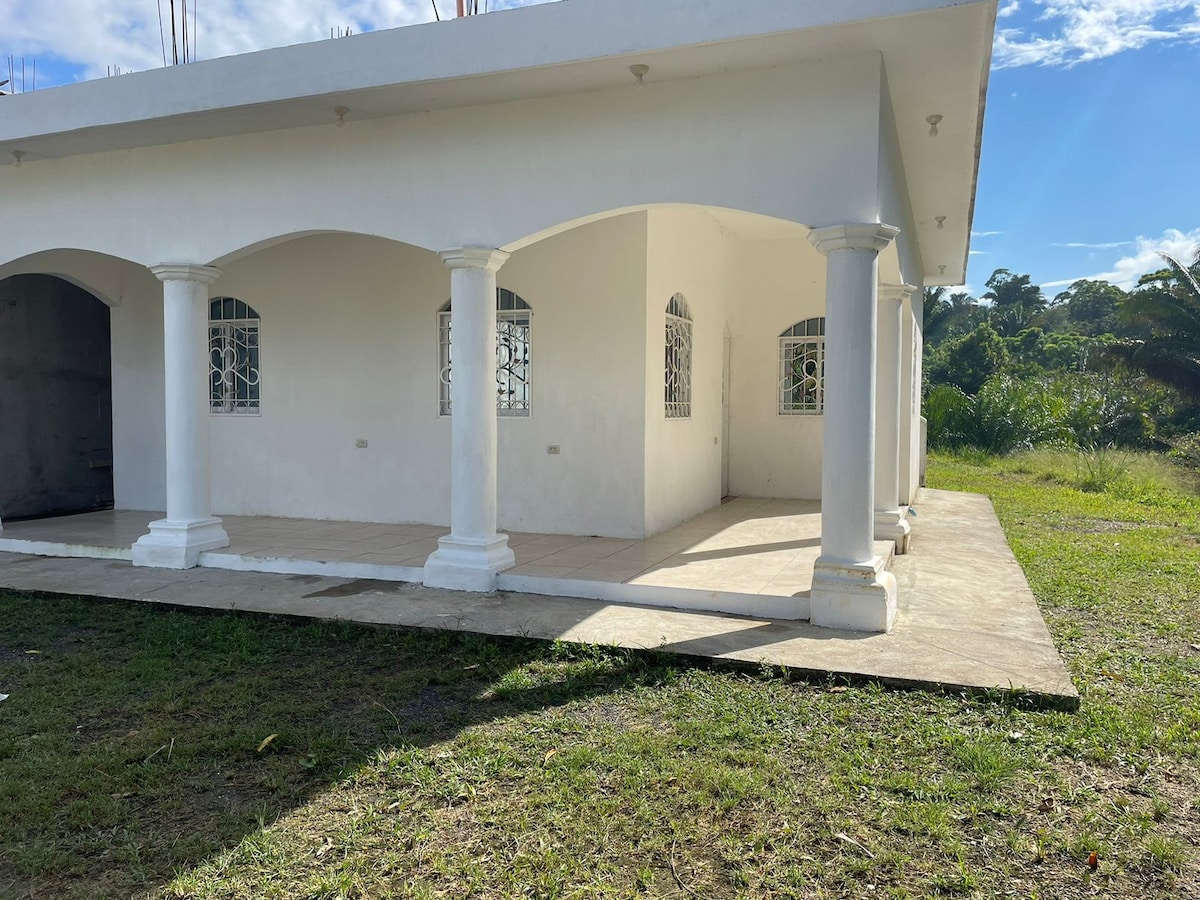 The exterior of the small house features a covered porch supported by white columns, providing shaded outdoor space. Large windows are highlighted by decorative metal grilles, allowing natural light to enter while maintaining privacy. A grassy area surrounds the building, enhancing the tranquil setting.
