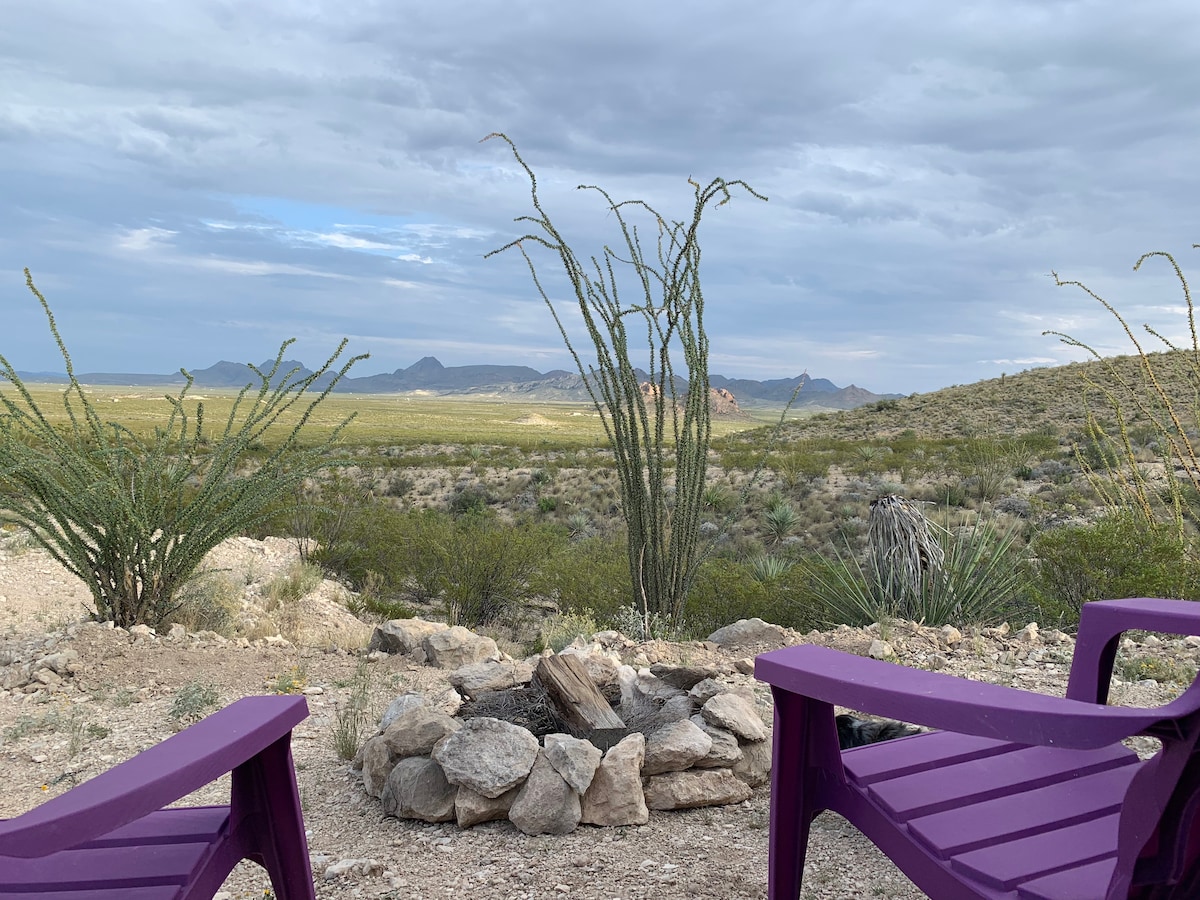 Two purple chairs frame a scenic view of the desert landscape, featuring a fire ring surrounded by a circle of stones. In the background, rolling hills and distant mountains extend under a mostly cloudy sky.