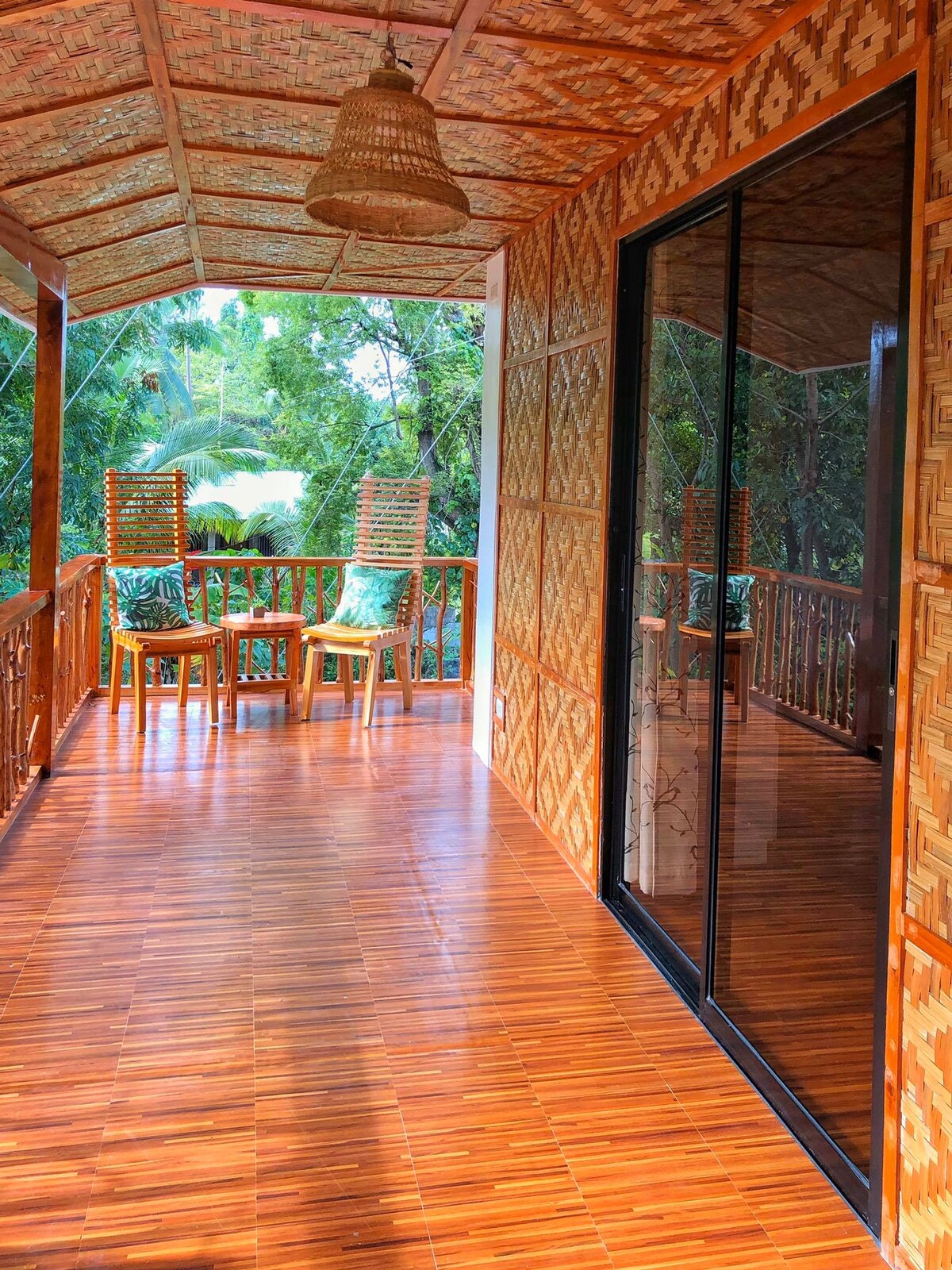 A shaded balcony area is adorned with wooden flooring and a textured bamboo ceiling. Two chairs are positioned at a small table, inviting relaxation. Large glass doors reflect the surrounding greenery, creating a harmonious connection with nature.