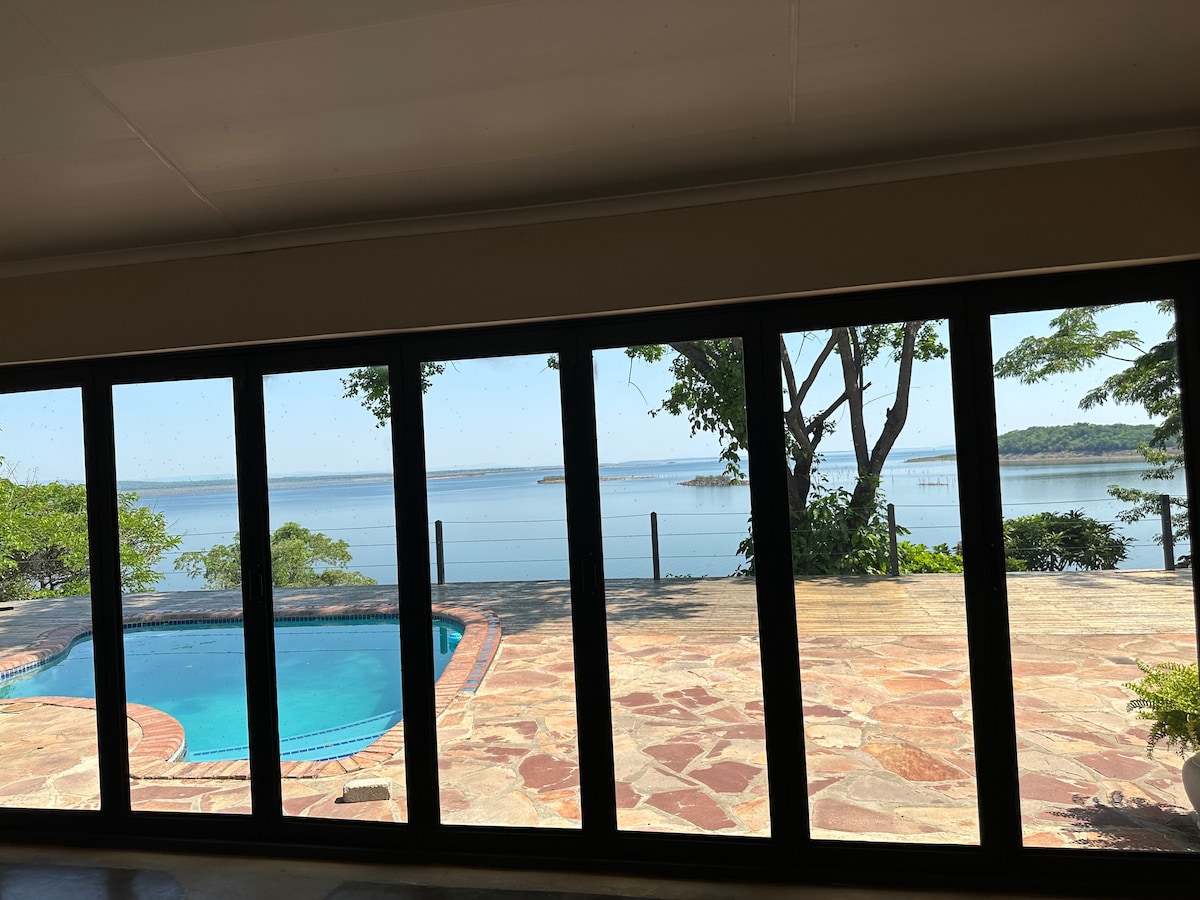 A panoramic view of the serene lake as seen through large sliding glass doors. The outdoor area features a swimming pool, while natural stones create a pathway leading towards the tranquil waters of the lake, framed by lush greenery.