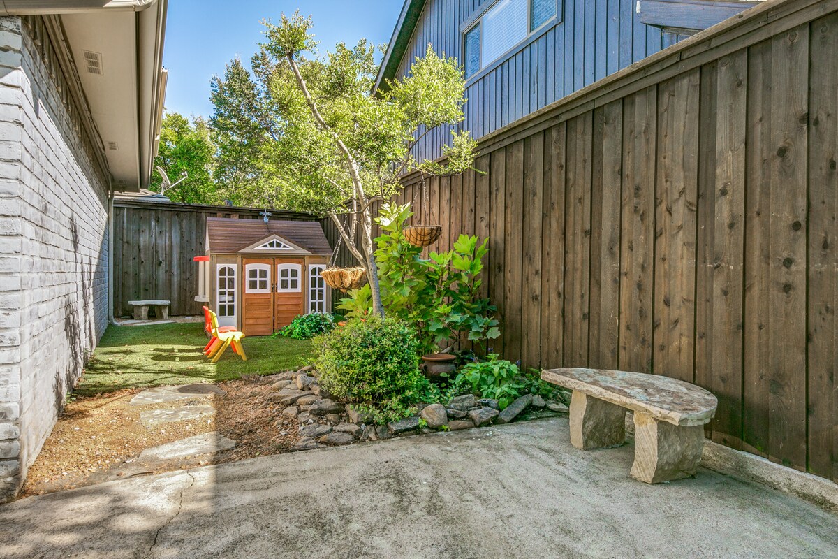 A private outdoor space features a well-maintained yard with grass and decorative stones. A small playhouse adds a playful touch, while a bench made of stone is positioned nearby. Lush greenery surrounds the area, enclosed by wooden fencing for privacy.