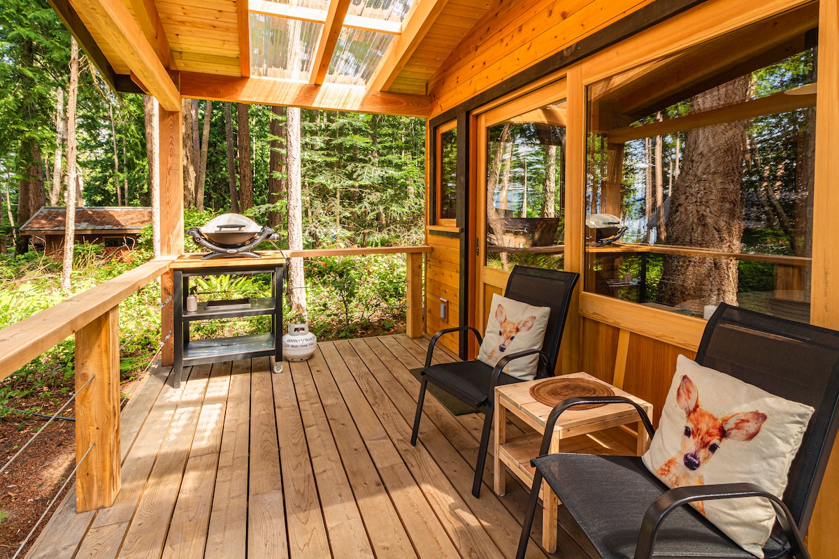 A semi-covered front deck features wooden flooring and is equipped with a Weber BBQ. Two comfortable chairs with decorative cushions are positioned around a small table, surrounded by trees that provide a serene forest setting.