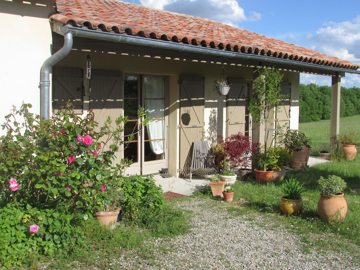 The exterior of the house features a welcoming entrance with a terracotta roof and wooden shutters. A gravel path leads to the door, which is surrounded by potted plants, including roses and greenery. Sunlight enhances the inviting atmosphere of the outdoor space.