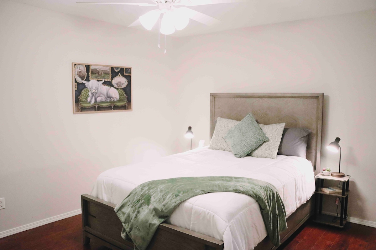 A cozy bedroom features a spacious bed adorned with white linens and a green throw blanket. Two bedside lamps provide soft lighting, while a framed artwork of a cow is displayed on the wall. A ceiling fan offers additional comfort.