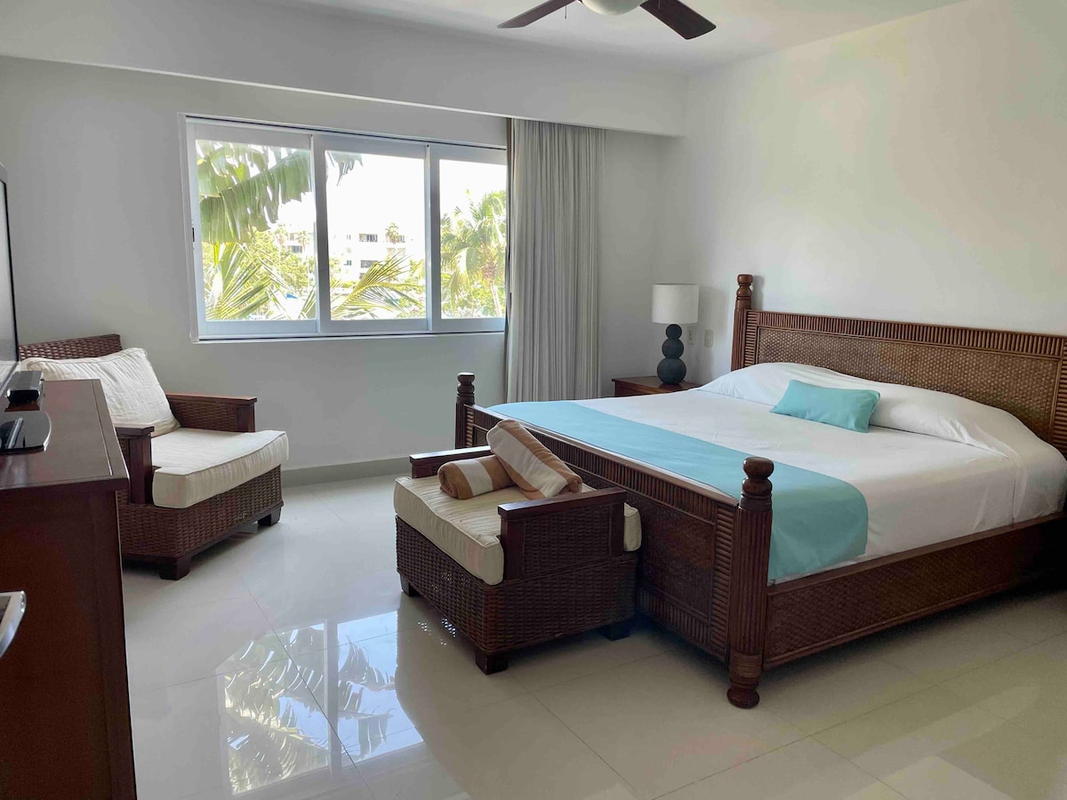 A spacious bedroom features a king-size bed with light bedding and a decorative throw. Two wicker chairs sit adjacent to the bed, complementing the wood furniture. Natural light pours through large windows lined with sheer curtains, offering a view of lush palm trees outside.