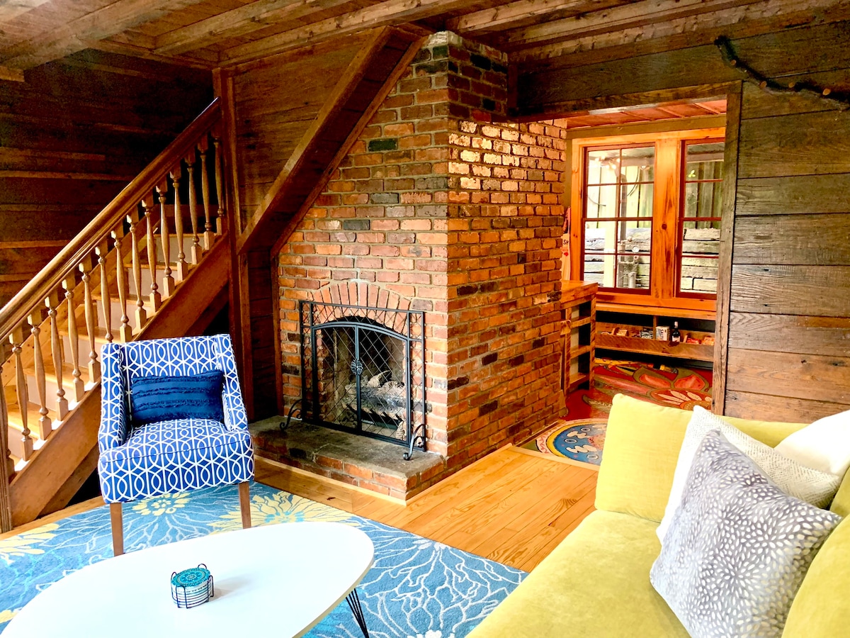 The living room features a cozy seating arrangement with a vibrant yellow couch and a blue patterned armchair. A rustic brick fireplace serves as a focal point, and wooden elements enhance the warm atmosphere. A staircase leads to the upper level, with natural light streaming through nearby windows.