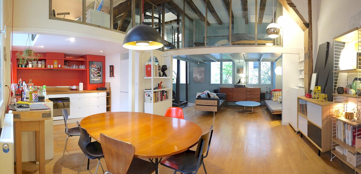 A spacious living area is characterized by wooden beams and large windows, allowing natural light to fill the room. A modern round dining table is surrounded by black and red chairs. The open kitchen features sleek cabinetry, while a comfortable sitting area includes a light gray sofa and a wooden coffee table.