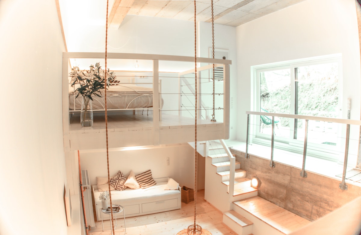 An open loft space features a suspended sleeping area above, framed by white railings. A spacious daybed is positioned below, complemented by natural light streaming through large windows. A staircase with a minimalistic design leads to the upper level, enhancing the airy atmosphere.