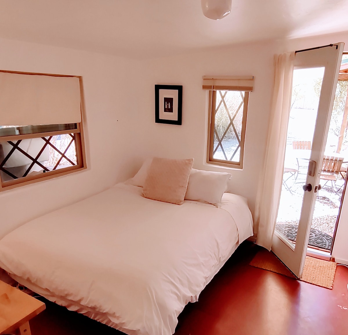 A bright sleeping area features a spacious bed covered with white bedding. Natural light enters through two small windows and a door that leads to a private outdoor patio with bistro seating. The room's walls are painted white, enhancing the airy atmosphere.